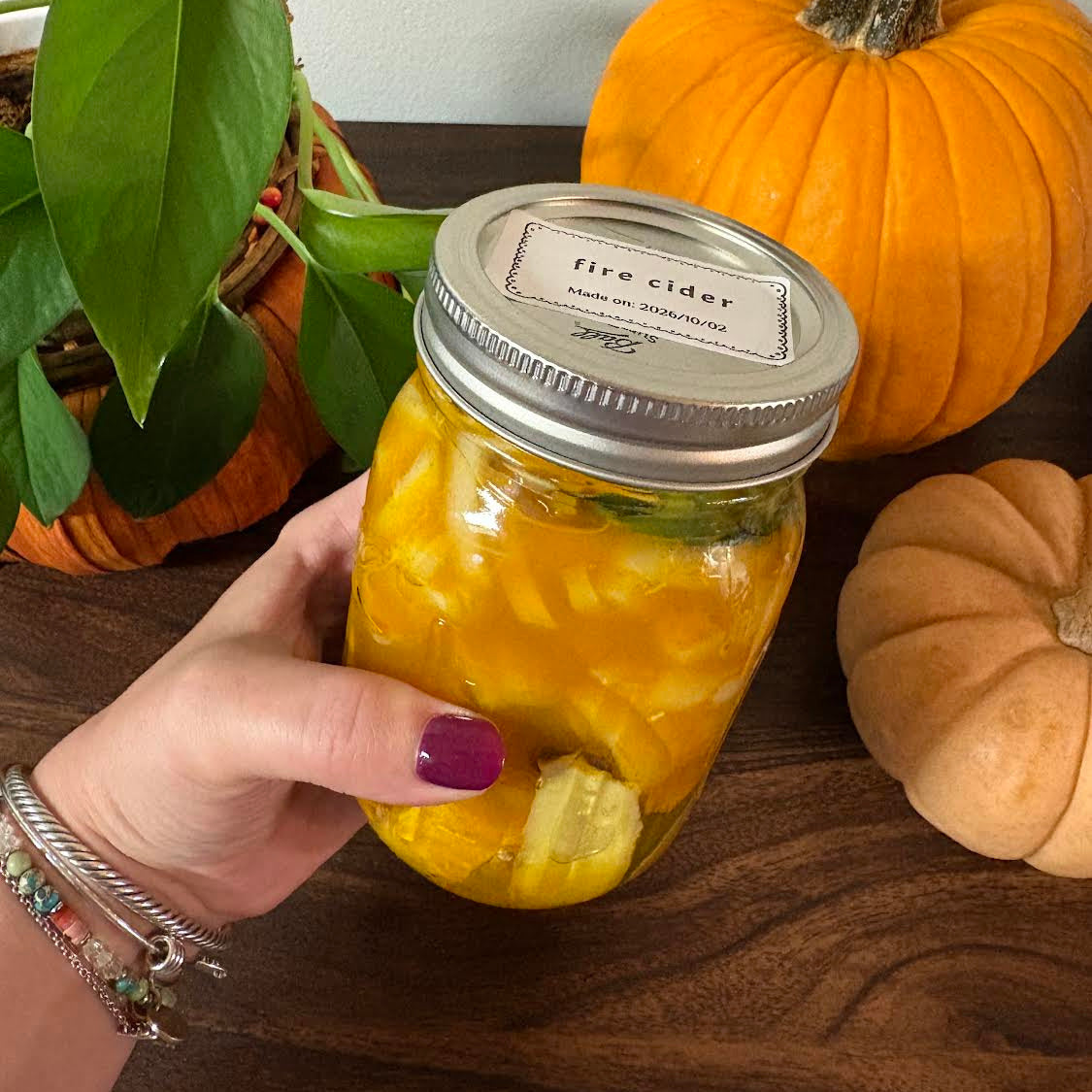 A hand with purple nail polish holds a glass mason jar filled with yellow fire cider, surrounded by pumpkins and a potted plant on a wooden table.