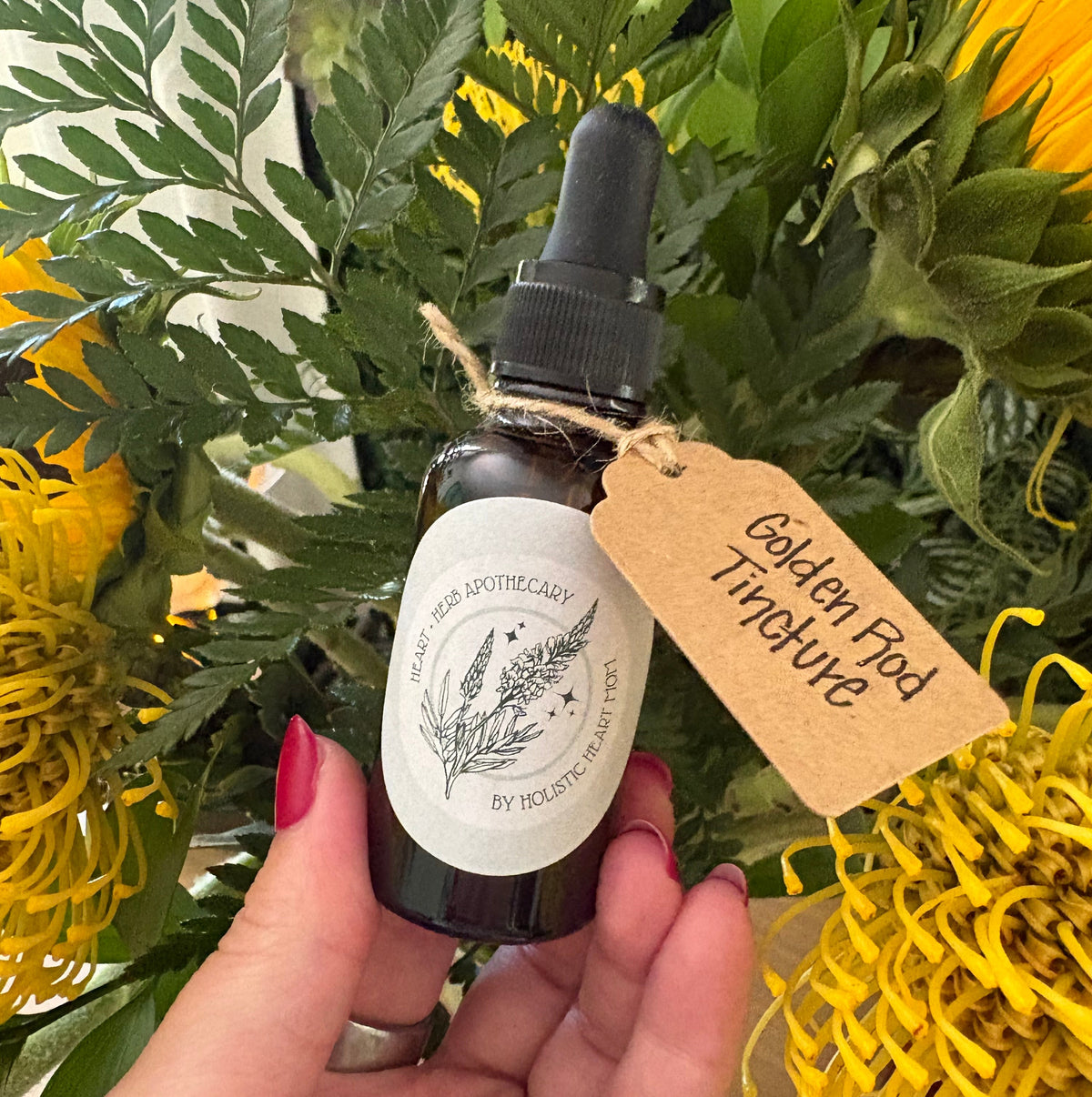A hand holds a small amber glass dropper bottle labeled 'Golden Rod Tincture' with a kraft paper hangtag, set against a background of green leaves and yellow flowers.