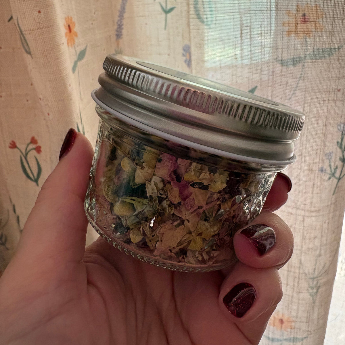 A hand with dark red nail polish holds a small clear glass jar filled with dried flowers and herbs, featuring a silver metal screw-on lid, set against a background of a floral-patterned curtain.