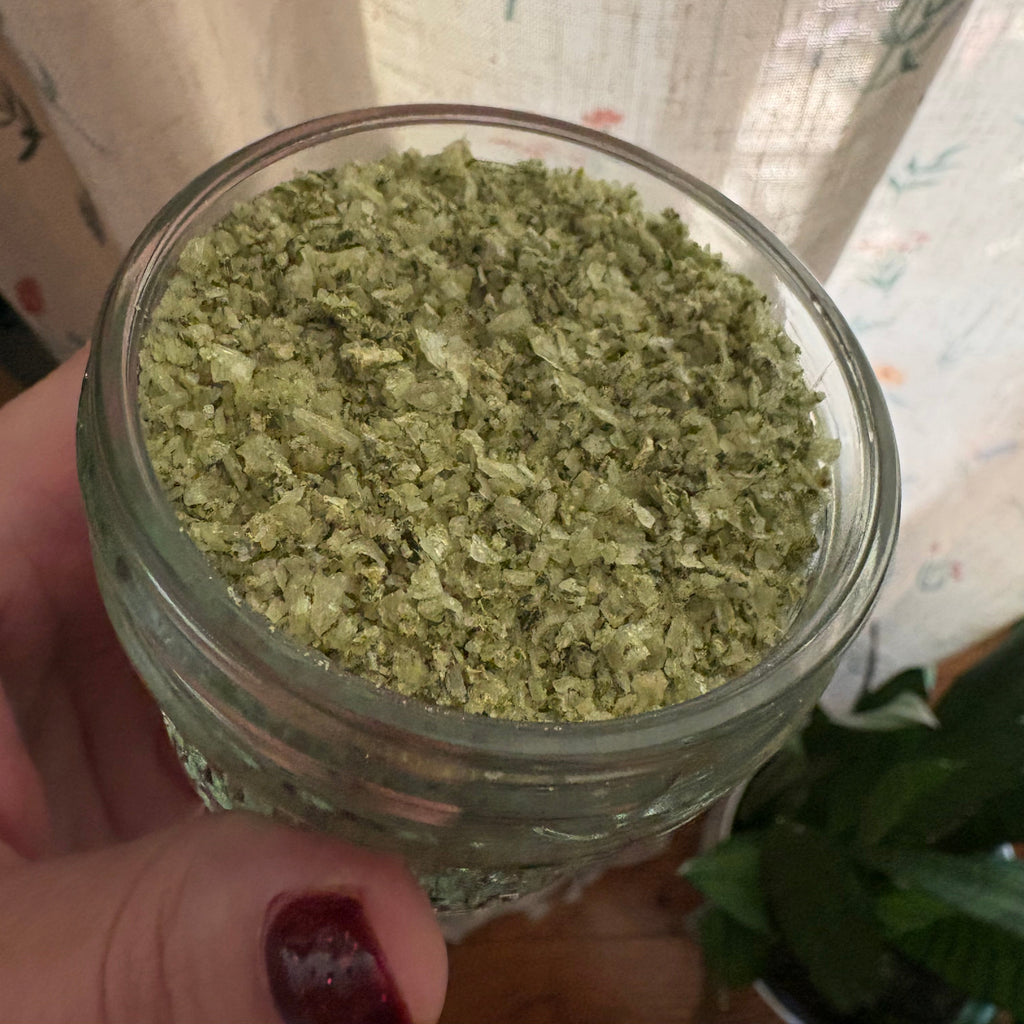 A hand with dark red nail polish holds a small clear glass jar filled with dried green herbal or botanical flakes, set against a background of patterned beige curtains and a green plant.