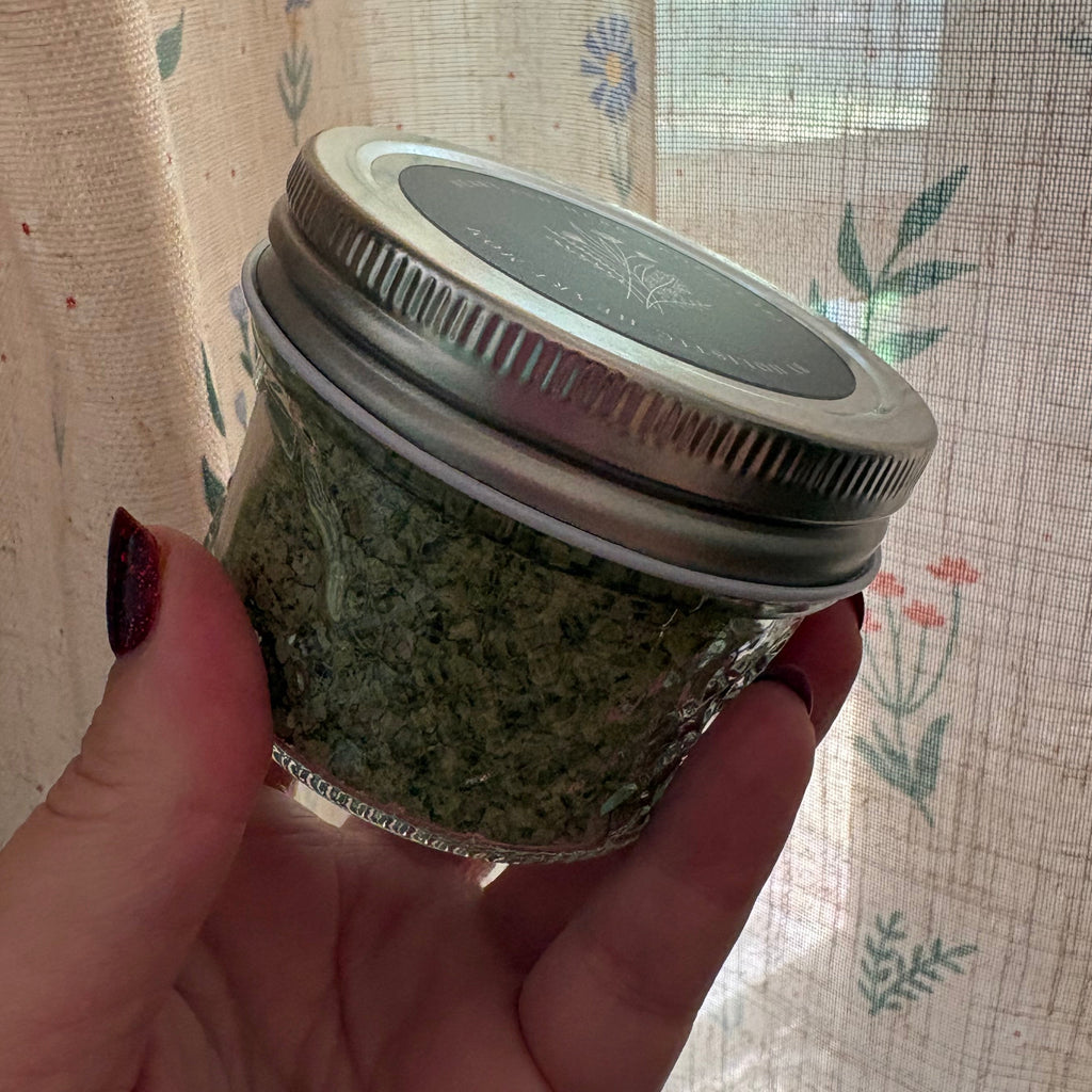 A hand holding a small glass jar filled with green herbal or herb paste, featuring a silver metal screw-on lid, set against a background of sheer floral-patterned curtains.