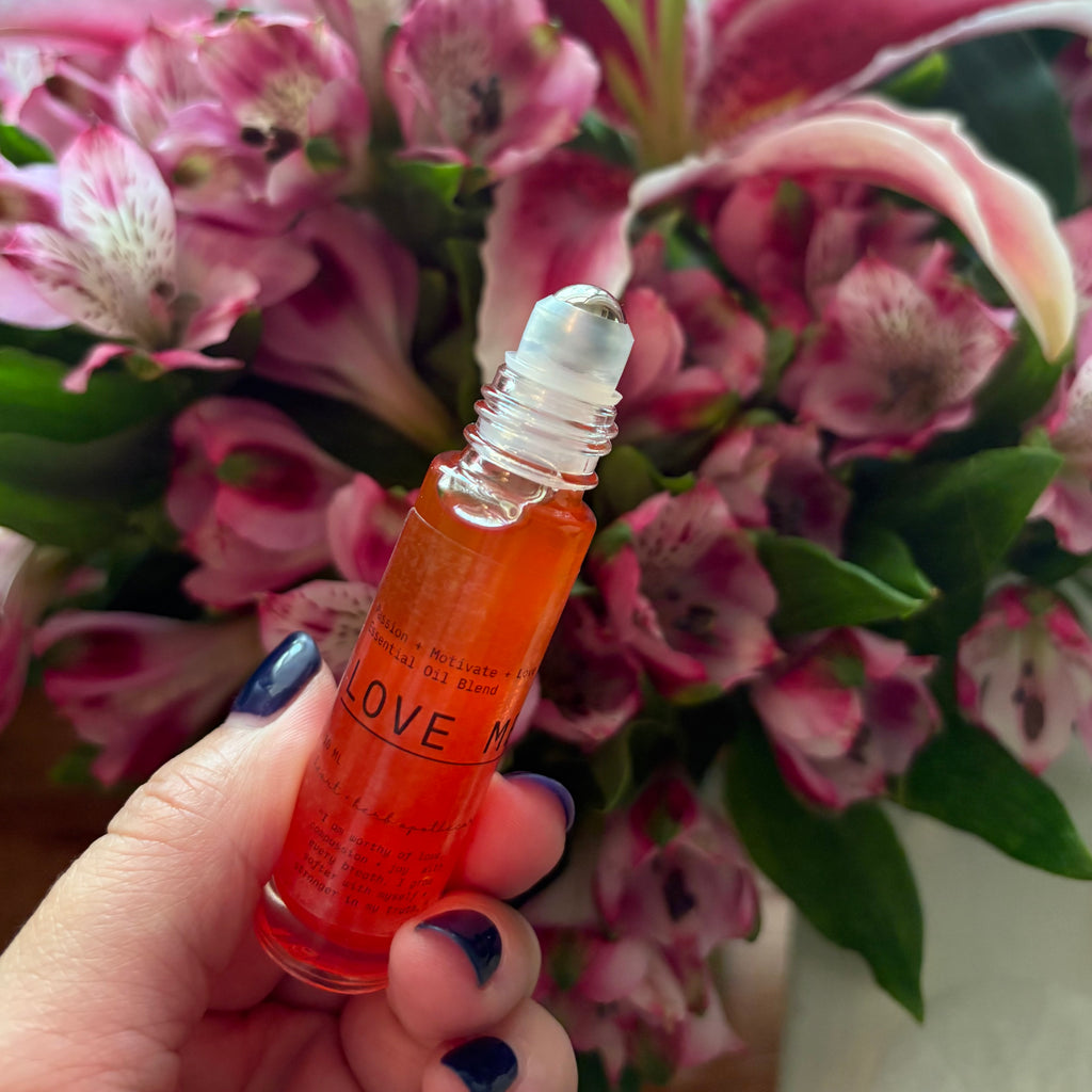 Hand holding a small orange bottle labeled 'LOVE' in front of pink flowers